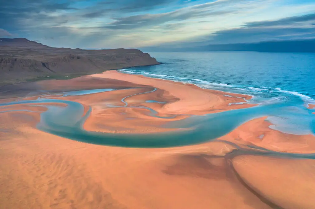 Aerial drone view of Raudasandur Beach in Iceland, showcasing the stunning interplay of azure water streams and vibrant yellow sand against a backdrop of dramatic cliffs.