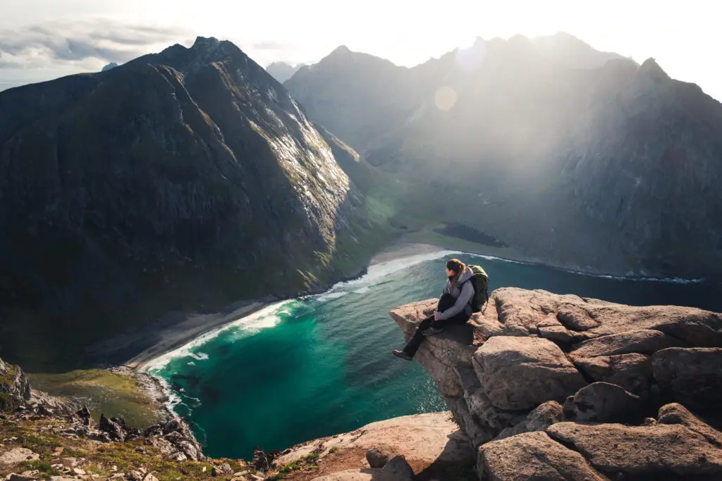 Woman sitting on the edge of Ryten mountain overlooking Kvalvika Beach in Lofoten Islands, Norway, showcasing a breathtaking scenic view of cliffs, beach, and blue ocean waters.