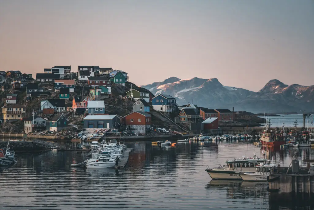 Sunset view of Maniitsoq, an Arctic city in Greenland, featuring colorful houses and stunning mountain backdrop during the midnight sun, with a panoramic view of the busy port including ships and boats.