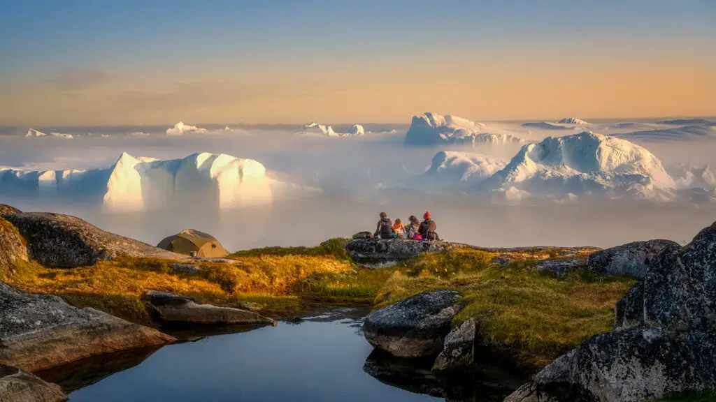 Tourists at Ilulissat Icefjord in Greenland enjoying a serene view of massive glaciers and a golden sunset from a rocky overlook with a camping tent nearby