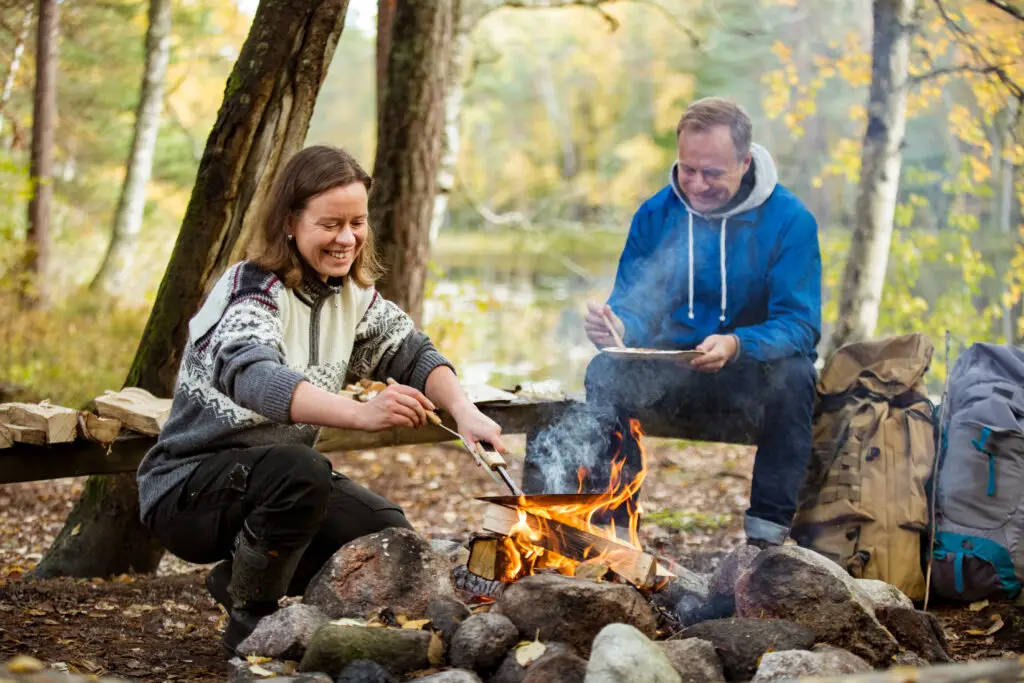 Man and woman enjoying outdoor cooking by a campfire in a Finnish forest, exemplifying a scenic and relaxing experience in Scandinavia's nature.