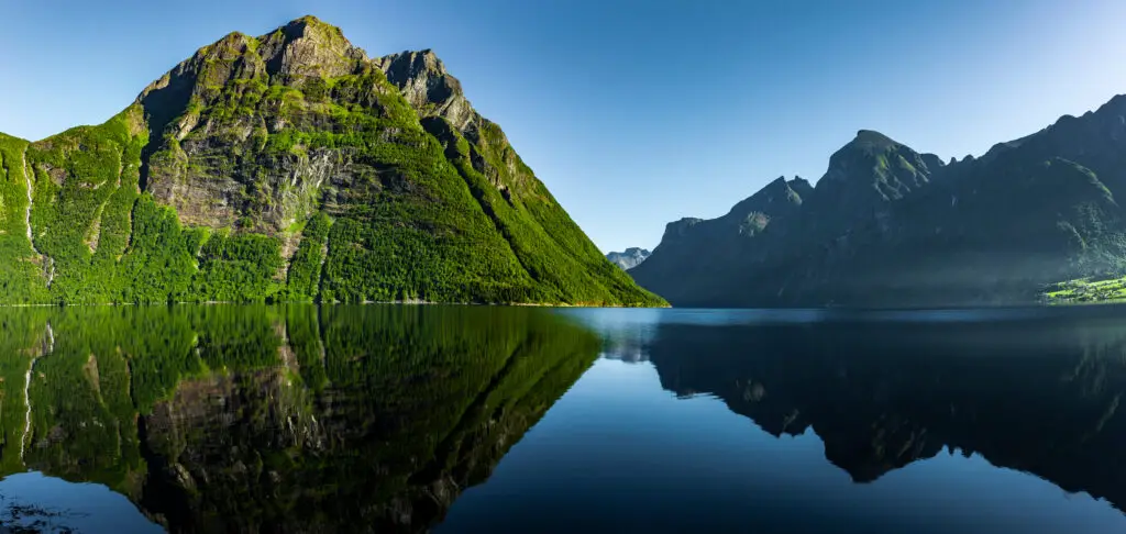 Panoramic view of Hjørundfjord in Norway displaying serene waters and towering green cliffs reflected perfectly in Scandinavia.