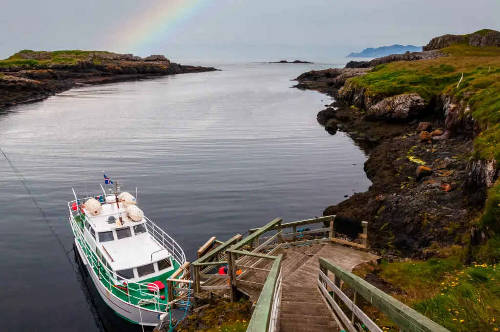 Rainbow over tranquil waters by a wooden pier and boat at Papey Island, Djupivogur, Iceland, highlighting scenic Nordic landscapes and tourist destinations.