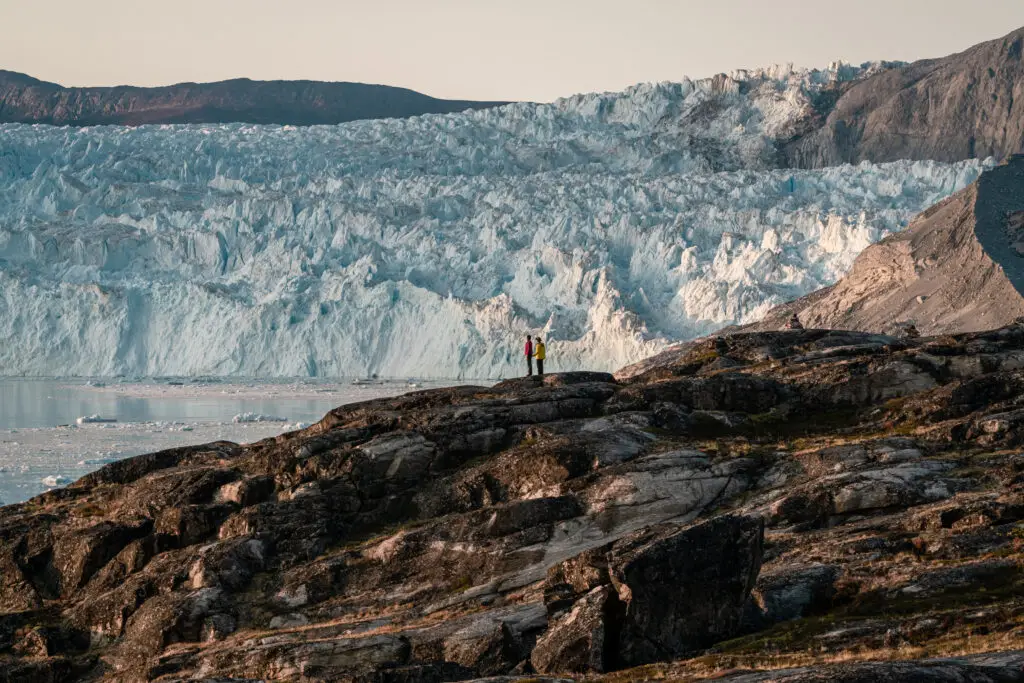 Tourists standing on rocky terrain admiring the vast Eqip Sermia Glacier in Greenland under the midnight sun, showcasing a dramatic ice calving event.