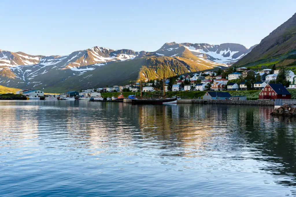 Scenic view of Siglufjordur, a small fishing town in a narrow fjord on the northern coast of Iceland, showcasing colorful houses and a serene harbor against a backdrop of majestic snow-capped mountains on a clear summer day.