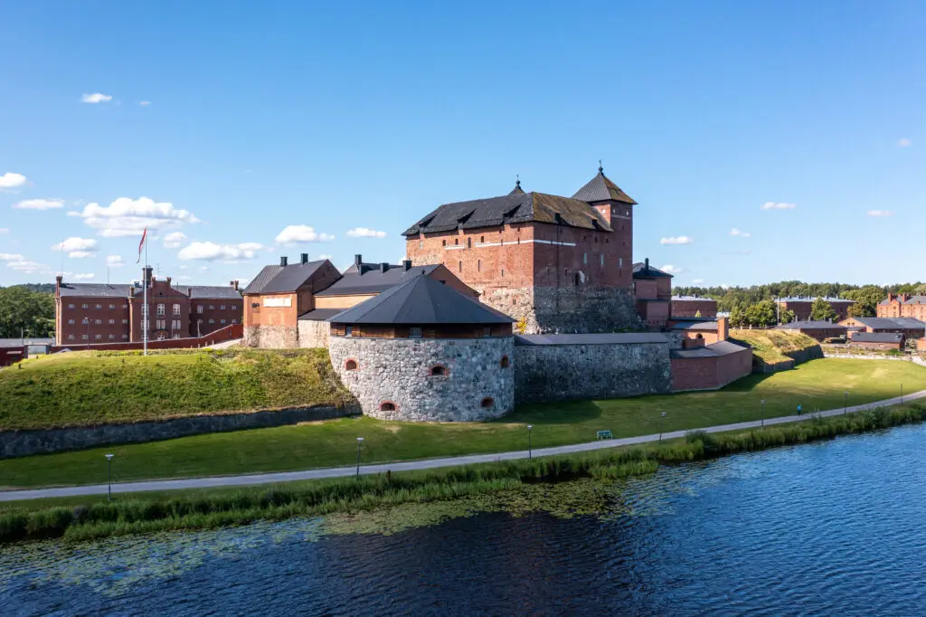 Summer view of Häme Castle, a prominent historical landmark in Hämeenlinna, Finland, beside a tranquil river under clear skies.