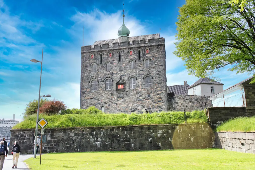 Tourists walking near the historic Bergenhus Fortress under a clear blue sky in Bergen, Norway.