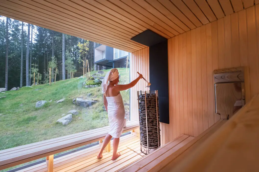 Woman in white towel enjoying a traditional Finnish sauna, pouring water on hot stones, with serene green forest visible through a large window, promoting relaxation and health in Finland.