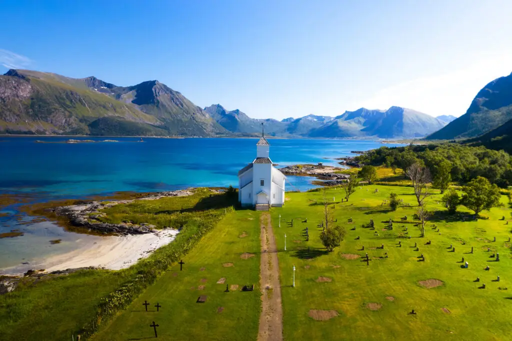 Aerial view of Gimsoy Church surrounded by vibrant green landscape and crystal blue waters in the Lofoten Islands, Norway, showcasing the stunning beauty of Scandinavian destinations.