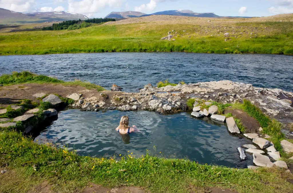 Woman relaxing in the Fosslaug hot pot, a natural geothermal hot spring in Iceland, surrounded by lush green hills and a flowing river under a clear blue sky.