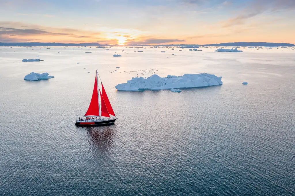 Sailboat with a red sail gliding through icy waters at sunset in Scandinavia, showcasing a serene and breathtaking Arctic tourism experience.
