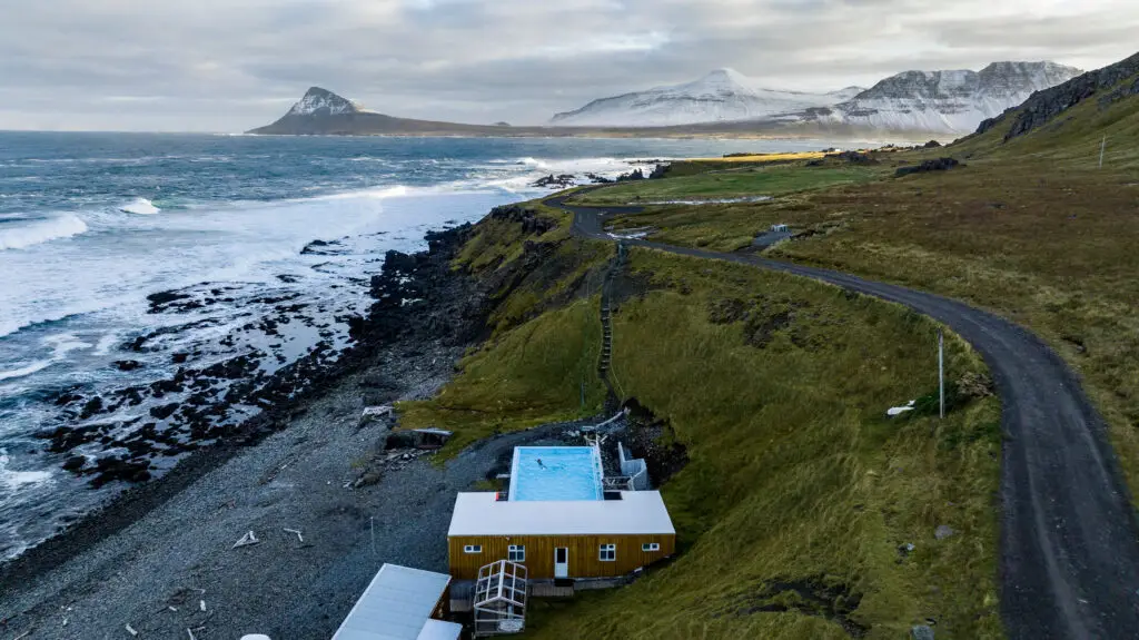 Aerial view of Krossneslaug geothermal pool on the Icelandic coast, featuring a swimmer, stormy seas, and fjords in the backdrop, surrounded by lush green grass.