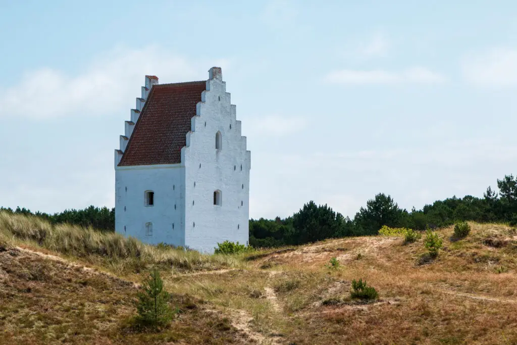 White historic Sand-Covered Church, Den Tilsandede Kirke, surrounded by grassy dunes under a blue sky in Denmark, a popular Scandinavian tourist attraction.