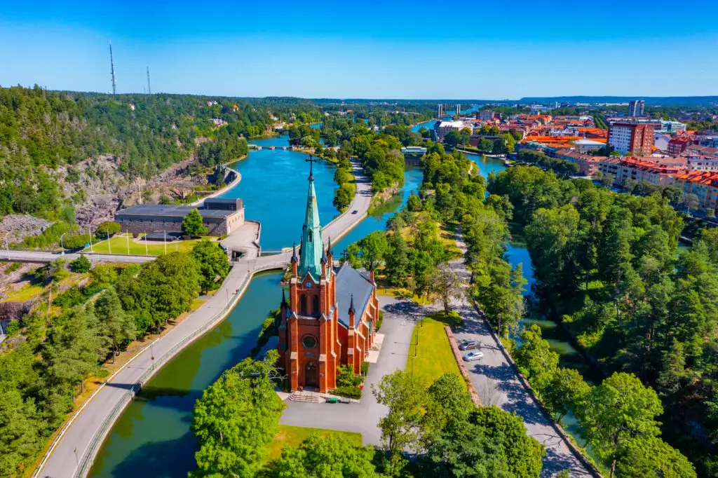 Aerial view of the historic red brick church in Trollhättan, Sweden, surrounded by lush greenery and flowing rivers, prominently showcasing Swedish architecture and natural beauty.