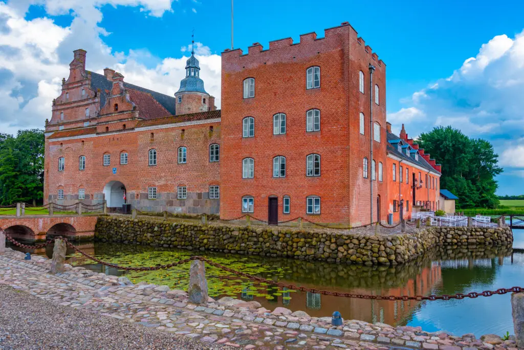 Broholm Slot Castle in Denmark, a historic red brick building surrounded by a moat and lush greenery, under a clear blue sky