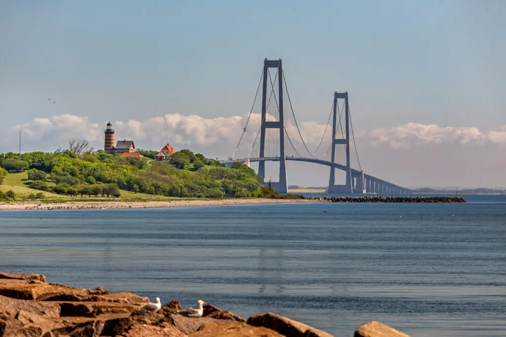 View of the Great Belt Bridge from Sprogø beach in Denmark, featuring a lighthouse and lush greenery, emphasizing Scandinavian tourism attractions.