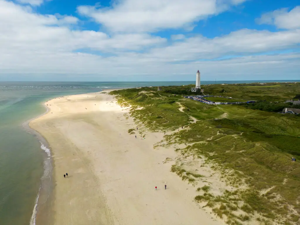 Aerial view of Blåvandshuk Fyr, the historic lighthouse in Blåvand, Denmark, overlooking a scenic sandy beach and lush greenery.
