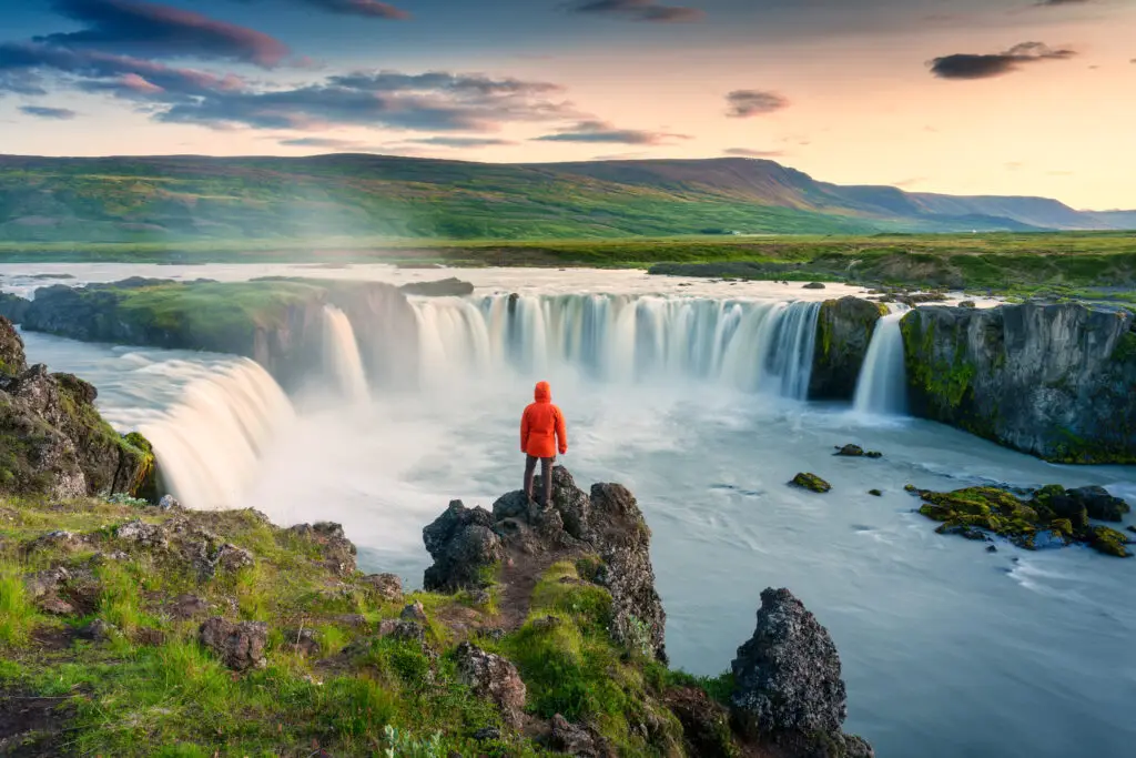 Male tourist in orange jacket standing on a cliff overlooking the majestic Godafoss waterfall in Iceland during a colorful summer sunset.