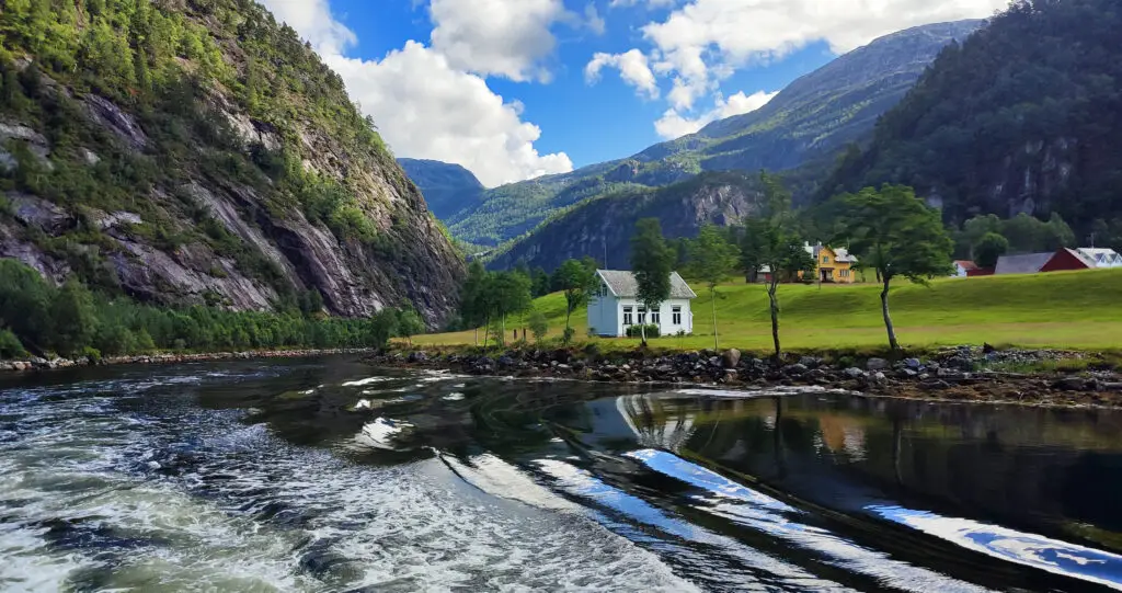 Scenic view from a Fjord Cruise in Mostraumen, Bergen, Norway, showing lush green cliffs and traditional Norwegian houses by the waterside, reflecting Norway's beautiful summer landscape.