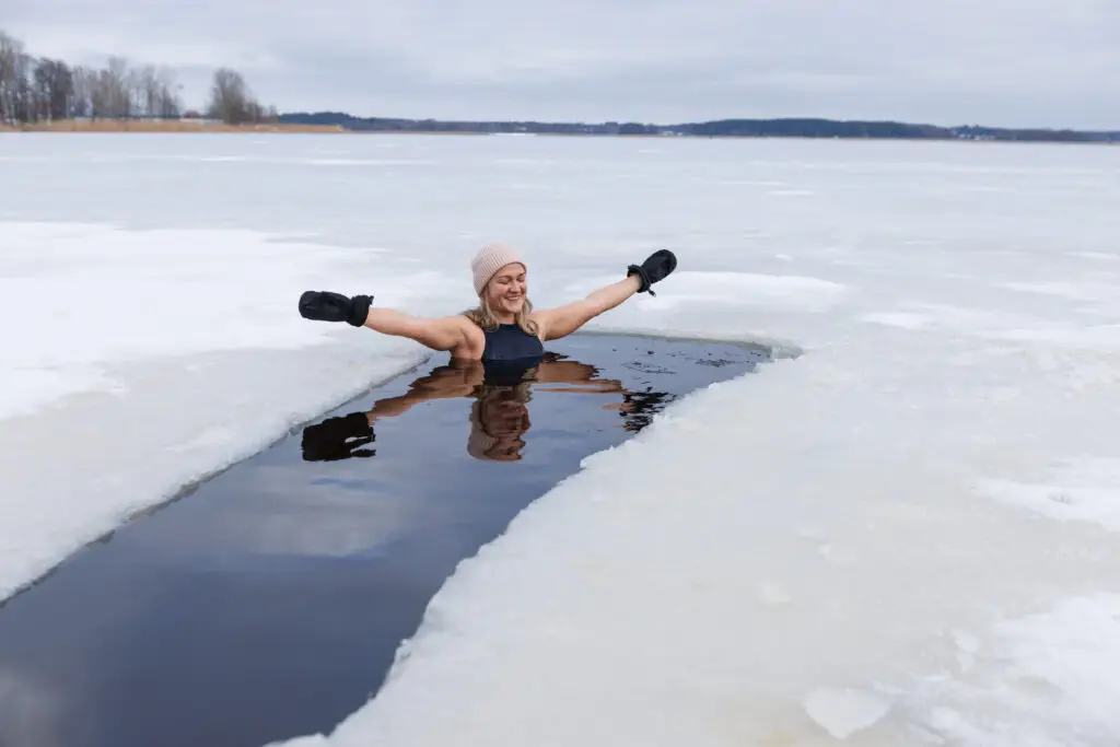 Young woman joyfully engages in winter swimming in a frozen lake in Scandinavia, demonstrating wellness and meditation practices in icy water, wearing a gray hat and gloves.