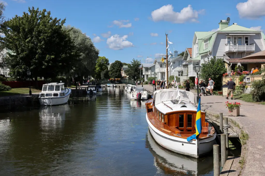 Pleasure boats moored along the calm Trosa River, surrounded by greenery and quaint houses in Trosa, Södermanland, Sweden, under a bright blue sky.